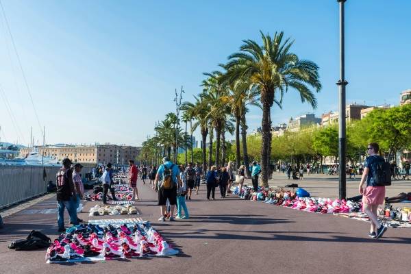 Langs med en havnepromenade har flere gateselgere spredd ut varene sine. I forkant av bildet står det en mann med mange rosa og hvite sko foran seg. Bak han står det en mann med mange hatter foran seg og videre bak dem står det mange andre som den med ulike varer. Mennesker spaserer bortover promenaden. På høyre side av bildet er det en rad med palmer. Himmelen er lyseblå.